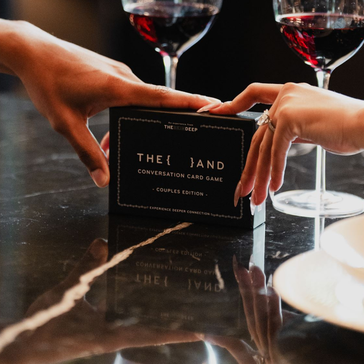 Two hands reaching towards a card game box titled 'The And' on a reflective surface with wine glasses in the background.